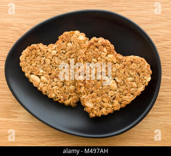 Zwei selbstgebackene herzförmige Erdnuss und Hafer flapjack Kekse für Valentines Tag auf ovalen schwarzen Teller gemacht auf hölzernen Tisch. Stockfoto