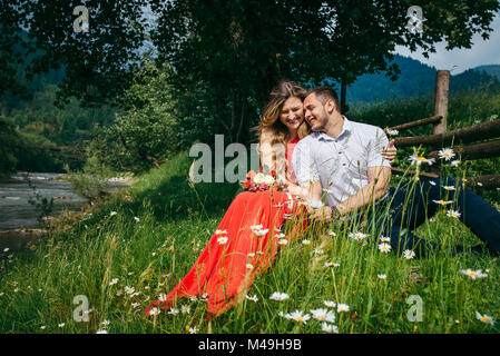 Glücklich lachend Paar ist zärtlich umarmen beim Sitzen auf der Kamille Wiese unter dem Baum in der Nähe von Fluss und Holzzaun. Die hübsche blonde Frau mit dem Bouquet. Stockfoto