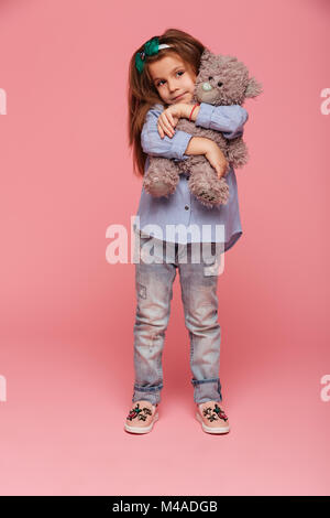In voller Länge Foto von adorable Kind Mädchen mit langen auburn Haar und legere Kleidung, ihren schönen Teddybär über rosa Hintergrund isoliert umarmen Stockfoto