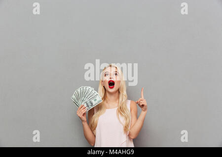 Portrait von eine aufgeregte Frau mit Bündel geld Banknoten und Zeigefinger über grauer Hintergrund Stockfoto