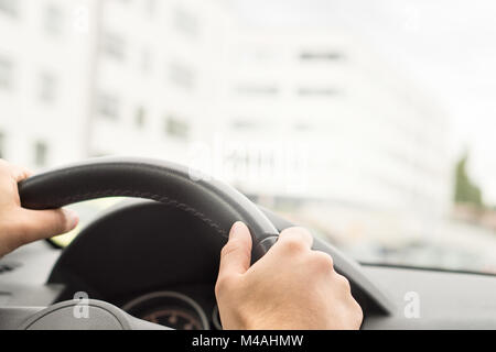 Man Autofahren in der Stadt. Fahrer mit Lenkrad mit beiden Händen. Reise, Reisen oder Pendeln Konzept. Gebäude in der verschwommenen Hintergrund. Stockfoto