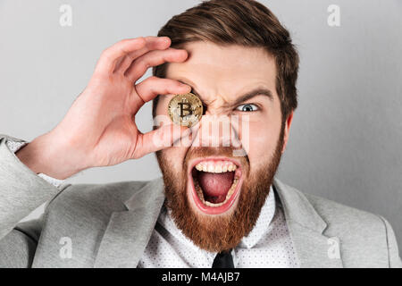 Nahaufnahme von einem schreienden Geschäftsmann in Anzug holding Bitcoin auf seinem Gesicht gekleidet über grauer Hintergrund Stockfoto