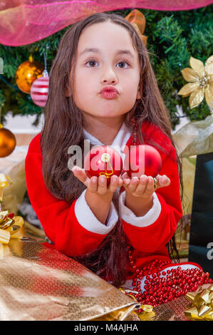 Nahaufnahme der glücklichen Mädchen mit einem roten Santa Kostüm und halten zwei Weihnachten Kugeln in den Händen tun lustiges Gesicht, mit einem Weihnachtsbaum hinter, Weihnachten Konzept Stockfoto