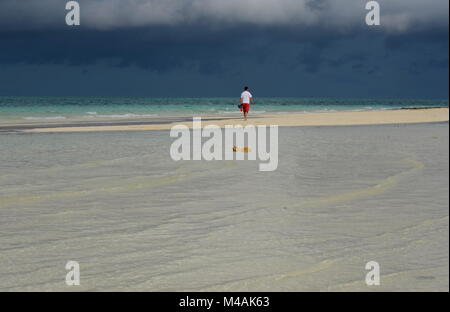 Eine einzelne kaukasischen Mann alleine zu Fuß in der Ferne auf eine leere tropischen Strand vor einem Sturm. Seascape der Gewitterwolken und das türkisfarbene Meer. Stockfoto