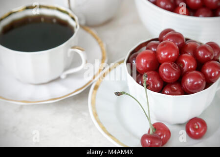 Auf dem Tisch liegt, ist eine Tasse Kaffee, Süßigkeiten, reifen Kirschen in einer Keramik Vase und eine Tasse. Stockfoto