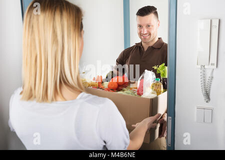 Lächelnden jungen Frau akzeptieren Karton voller Lebensmittel aus Delivery Man Stockfoto