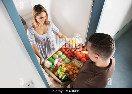 Ansicht von Lächelnden jungen Frau akzeptieren Karton voller Lebensmittel aus Delivery Man Stockfoto