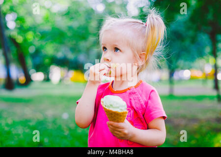 Wenig lustig Mädchen blond Süßigkeiten blaue Eis in der Waffel Schale auf einem grünen Hintergrund im Park. verschmierte Gesicht und Wangen und lacht. In hellen stilvollen Kleidung Stockfoto