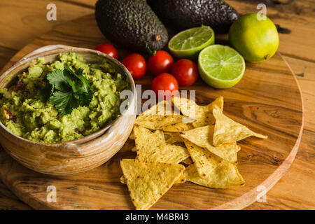 Schüssel mit Guacamole Dip mit avocados Tortilla Chips Kirschtomaten und Limetten auf einem Schneidebrett Stockfoto