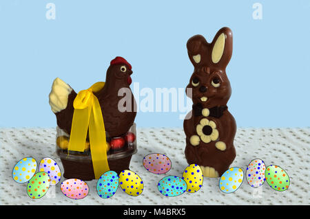 Eine Henne auf einem Korb und eine Schokolade Kaninchen mit farbigen Eier auf eine Tischdecke und blauem Hintergrund. Stockfoto