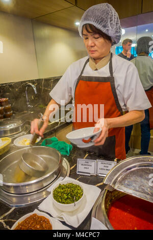 Singapur, Singapur - Januar 30. 2018: Nicht identifizierte Frau mit einem riesigen metallenen Löffel servieren die Penang asam laksa Reis Nudeln innerhalb der weißen Schale mit Würziger Fisch tamarind Suppe und frischen Kräutern in Singapur Stockfoto