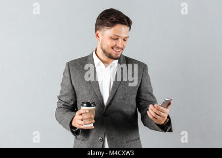 Porträt eines lächelnden Geschäftsmann mit Mobiltelefon suchen, während Sie Kaffee über grauen Hintergrund gehen Stockfoto
