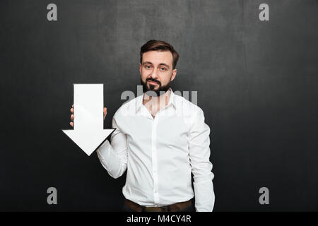 Bild von pessimistisch umgekippt Mann 30 s Holding leere Rede Pfeil abwärts Regie über dunkle graue Wand Platz kopieren Stockfoto