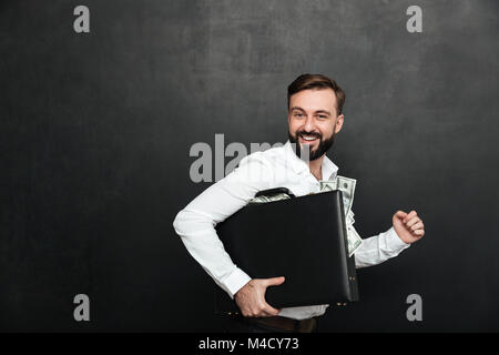 Lustiges Bild der glücklichen Mann mit schwarzen Aktenkoffer voller Dollar Banknoten im Inneren über dunkelgrauen Hintergrund isoliert Stockfoto