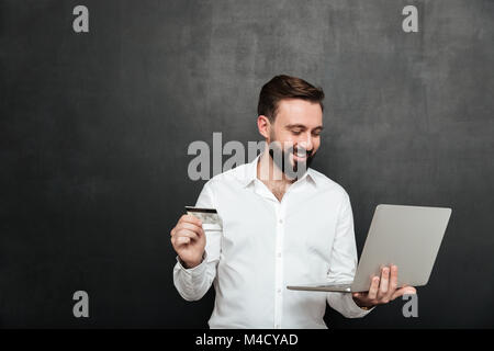 Portrait von fröhlichen Menschen tun online Zahlung im Internet mit Notebook und Kreditkarte über dunkelgrauen Hintergrund isoliert Stockfoto