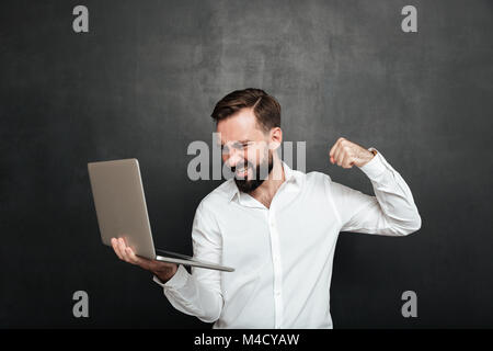 Portrait von aggressiven bärtigen Mann mit Silber personal computer und werfen die Punch In-Bildschirm über die dunkelgraue Wand isoliert Stockfoto