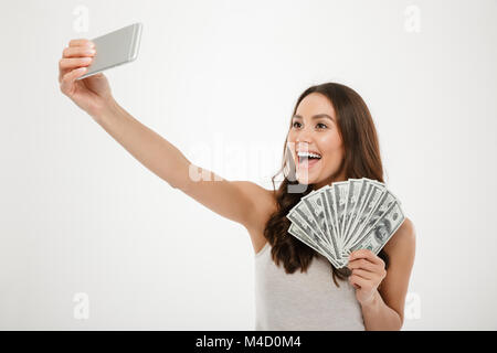 Foto von Lucky reiche Frau, die selfie auf Silber Mobiltelefon halten Sie viel Geld Dollarscheine über weiße Wand isoliert Stockfoto