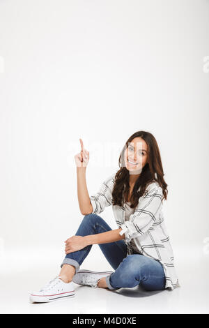 Full-length Portrait von gerne Frau in legere Kleidung auf dem Boden saß und nach Index Finger mit einem breiten Lächeln über weiße Wand isoliert Stockfoto