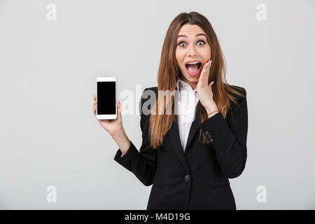 Schöne Dame in offiziellen Anzug, die Kamera mit geöffnetem Mund und zeigt Smartphone mit leerer Bildschirm isoliert Stockfoto