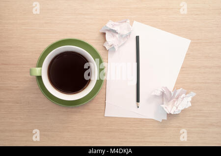 Eine grüne Tasse Kaffee auf dem Hintergrund einer Tabelle und einem weißen leeren Blatt Papier, Bleistift und zerknitterten Papierstücke simuliert Nervosität, psyc Stockfoto