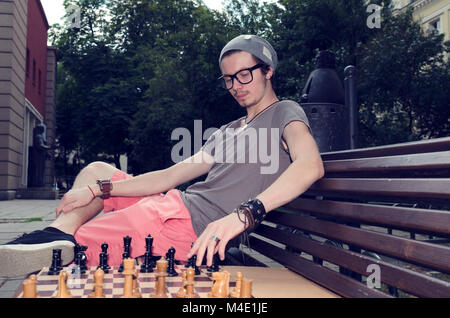 Junger Mann spielt Schach im Park. Öffentlicher Park. Öffentliche Gebäude. Stockfoto