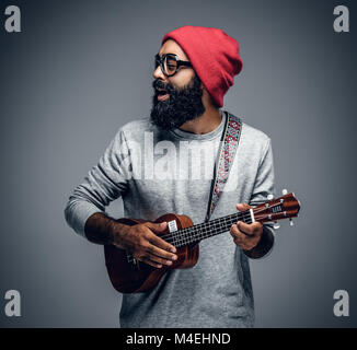 Portrait eines bärtigen Hipster männlich in Red Hat das Spielen auf Ukulele. Auf grauem Hintergrund. Stockfoto