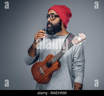 Portrait eines bärtigen Hipster männlich in Red Hat das Spielen auf Ukulele. Auf grauem Hintergrund. Stockfoto