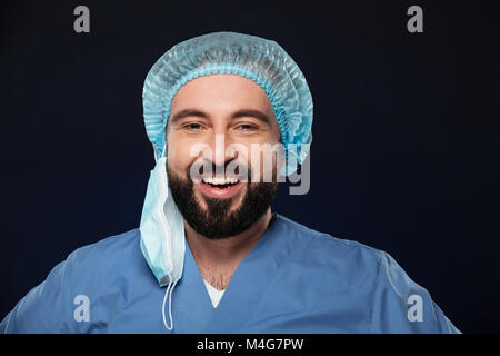 Nahaufnahme, Porträt eines lächelnden bärtige männliche Chirurg trägt Gesichtsmaske und sterilen hat über dunklen Hintergrund isoliert Stockfoto