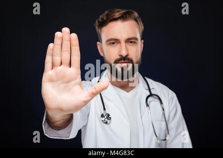 Porträt einer zuversichtlich männlicher Arzt in Uniform mit Stethoskop mit stop Geste mit Palm über dunklen Hintergrund gekleidet Stockfoto