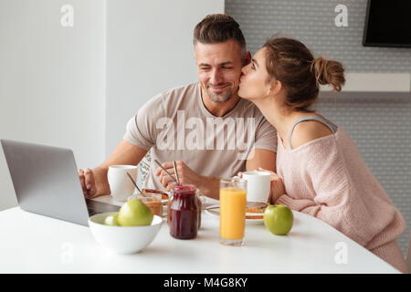 Portrait einer jungen Liebespaar, das Frühstück am Tisch in einer Küche zu Hause sitzen mit Laptop und küssen Stockfoto