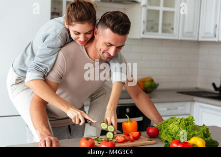 Porträt eines lächelnden Liebespaar kochen Salat zusammen und stehen auf einer Küche zu Hause, Frau sitzt auf der Schulter des Mannes und Schneiden von Gemüse Stockfoto
