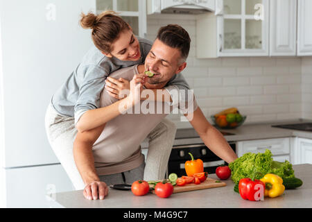 Porträt eines glücklich verliebten kochen Salat zusammen, während auf einer Küche zu Hause stehen, Frau sitzt auf der Schulter des Mannes und Schneiden von Gemüse Stockfoto