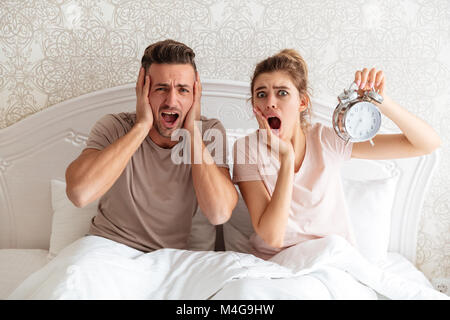 Paar schöne sitzen zusammen auf dem Bett mit Wecker und in die Kamera zu Hause Geschockt Stockfoto