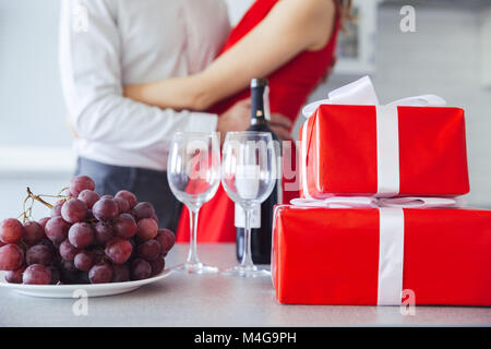 Nahaufnahme der hübschen Geschenkboxen, Flasche Wein und Traubenmost auf Tisch. Liebhaber umarmt im Hintergrund Stockfoto