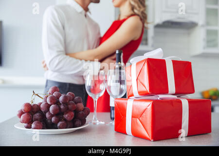 Nahaufnahme der hübschen Geschenkboxen, Flasche Wein und Traubenmost auf Tisch. Liebhaber umarmt im Hintergrund Stockfoto