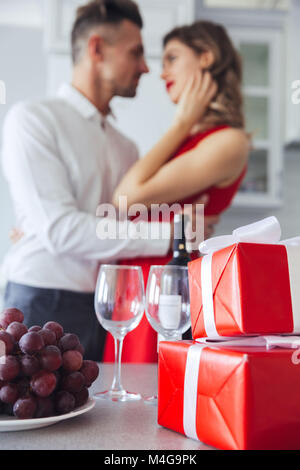 Nahaufnahme der vertikalen hübschen Geschenkboxen, Flasche Wein und Traubenmost auf Tisch. Liebhaber umarmt im Hintergrund Stockfoto