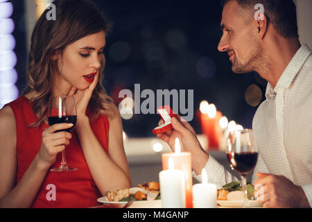 Schockiert Frau im roten Kleid holding Glas Wein und ihr Mann, der ihr Box mit Verlobungsring während romantische Abendessen Stockfoto