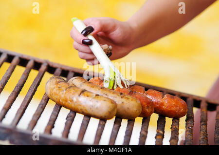 Nahaufnahme von Frau s Hand eine lange weiße Zwiebel Öl hinzufügen zu einem grillen Würstchen am Grill zubereitet. Grillplatz im Garten. Bayerische Wurst Stockfoto