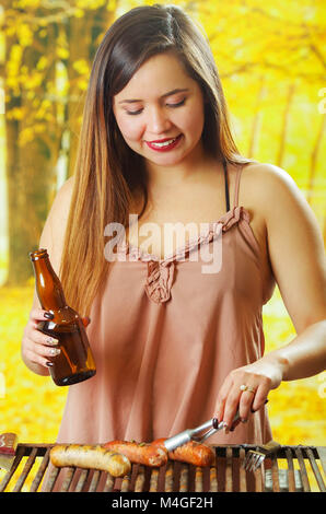 Nahaufnahme von lächelnd schöne junge Frau mit einer Flasche in der einen Hand und mit einer Zange, und drehen Sie das grillen Würstchen am Grill zubereitet. BBQ. Bayerische Wurst Stockfoto