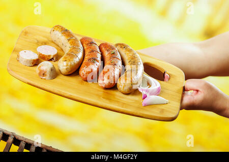 Nahaufnahme von Frau halten in ihren Händen ein gegrilltes Würstchen auf Holz Schneidebrett, Grill im Garten. Bayerische Wurst, in einem verschwommenen Hintergrund Stockfoto