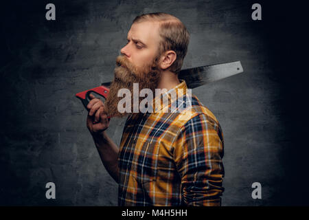 Studio Portrait von bärtigen Zimmermann in einem karierten Hemd hält Handsäge auf Grau vignette Hintergrund. Stockfoto