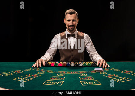 Porträt von einem Croupier hält Spielkarten, Casino Chips auf dem Tisch. Schwarzen Hintergrund. Ein junges Männchen croupier in einem Hemd, Weste und Fliege ist Stockfoto