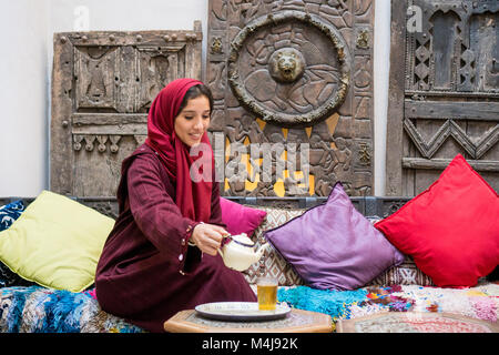 Arabische Frau im traditionellen roten Kleidung mit Hijab auf ihrem Kopf gießt ein Kaffee in der traditionellen orientalischen Ambiente Stockfoto