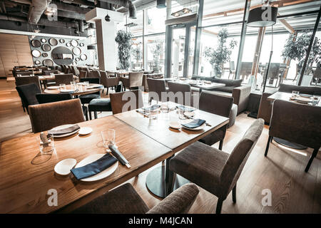 Spektakuläre Inneneinrichtung des modernen Restaurant Im eleganten Grautönen. Die Haupthalle mit den hölzernen Tische, Stühle und Samtsofas. Glaswand Stockfoto