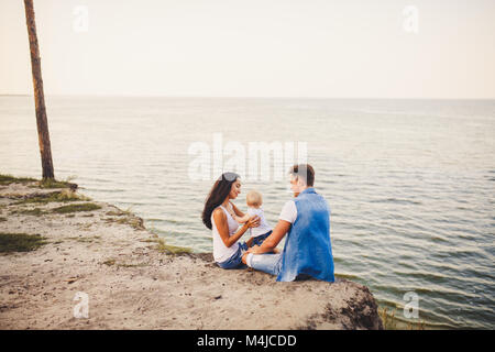 Thema Familienurlaub mit kleinen Kind auf die Natur und das Meer. Mama, Papa und Tochter von einem Jahr sitzen in Umarmung, Mädchen in die Hände des Menschen, Witz Stockfoto