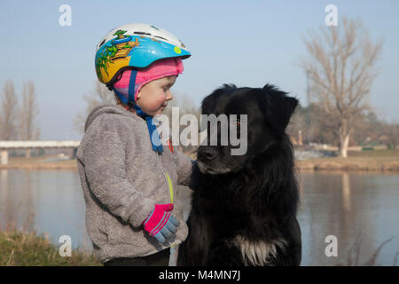 Kleine 3 Jahre alte Mädchen mit dem Fahrrad Helm neben ihr schwarz haarige Hund neben dem Fluß in der kalten Winter Zeit steht. Stockfoto