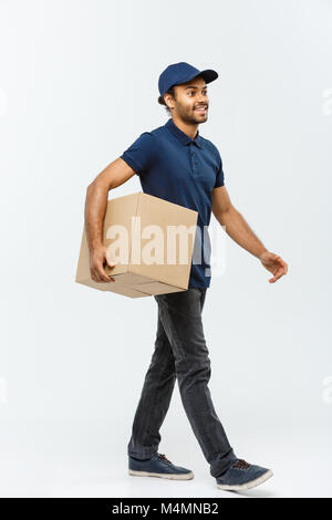 Lieferung Konzept - Portrait von Happy African American Lieferung Mann in blauem Stoff zu Fuß eine Box Paket an den Kunden zu senden. Auf Grau studio Hintergrund isoliert. Kopieren Sie Platz. Stockfoto