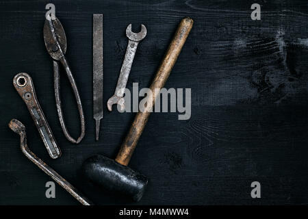 Alte, rostige Werkzeuge liegen auf einem schwarzen Holztisch. Hammer, Meißel, Metall, Schere, Schraubenschlüssel. Ansicht von oben. Kopieren Sie Platz. Immer noch leben. Flach Stockfoto