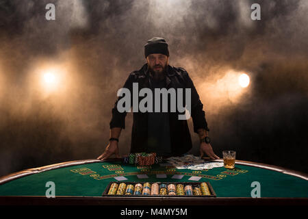 Männliche Spieler Poker spielen, Rauch dunkle Farbe Intensität. Ein großer bärtiger Mann mit einem Glas Whiskey in der Hand steigt über den Poker Tisch Stockfoto