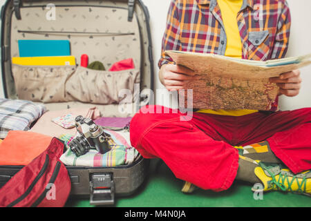 Ein Mann plant eine Reise mit einer Karte in den Händen, während er in der Nähe eines Koffers mit Dingen sitzt. Stockfoto